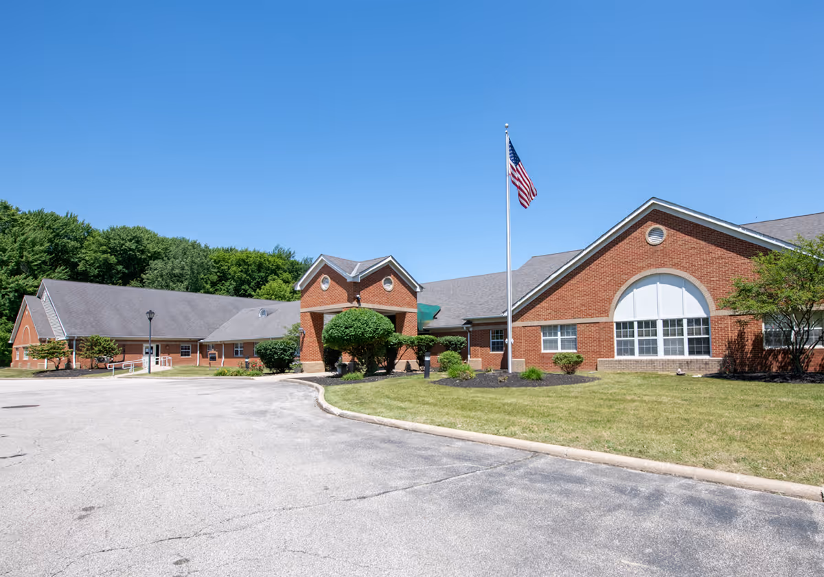 Exterior view of Hopkins Rehabilitation and Care Center, a single-story brick building with a gray roof, an American flag on a flagpole, and neatly trimmed bushes and grass under a clear blue sky.