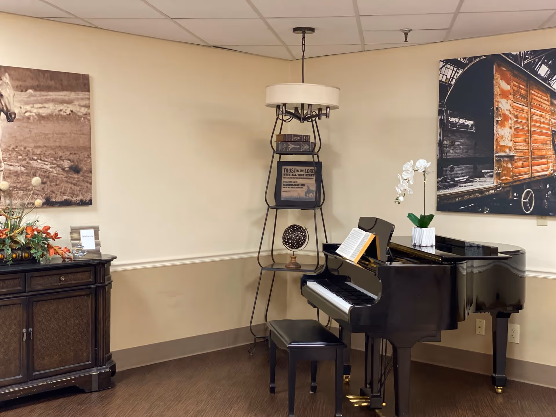 Interior corner of a room featuring a black grand piano with an open music book and a white orchid plant on top. To the left is a dark wooden cabinet with a floral arrangement and a framed photo. Behind the piano is a tall metal shelf with decorative items and a lamp on top. The walls are beige with two large framed pictures, one of a horse and the other of a rustic wooden train car.
