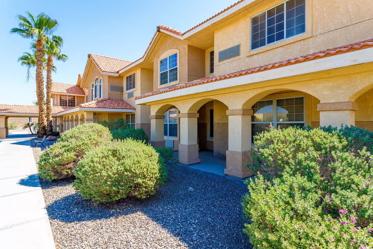 Front exterior of a two-story stucco senior living building with arched walkways, desert landscaping, and palm trees.