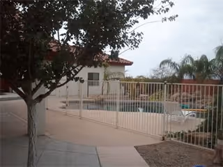 Gated outdoor pool area with lounge chairs, a tree, and a nearby single-story building under an overcast sky.