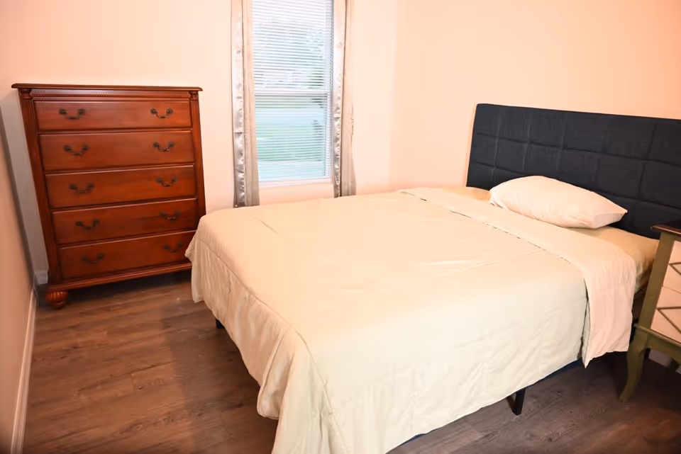 A neatly made bedroom with a bed, dresser, nightstand, and window.