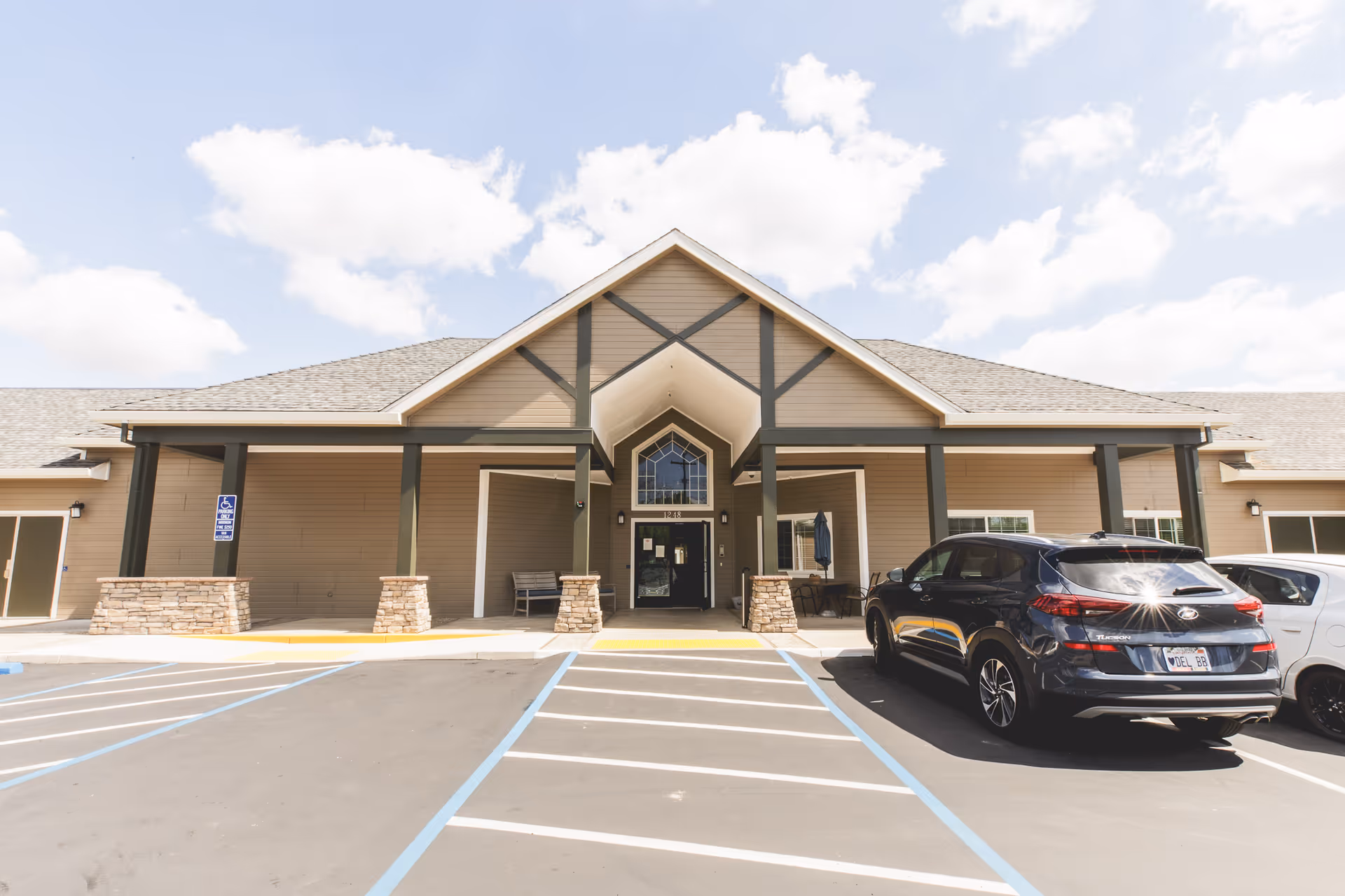 Front exterior view of Orangeburg Manor building with a covered entrance supported by stone pillars and dark beams. There are two cars parked in front, and the sky is partly cloudy.