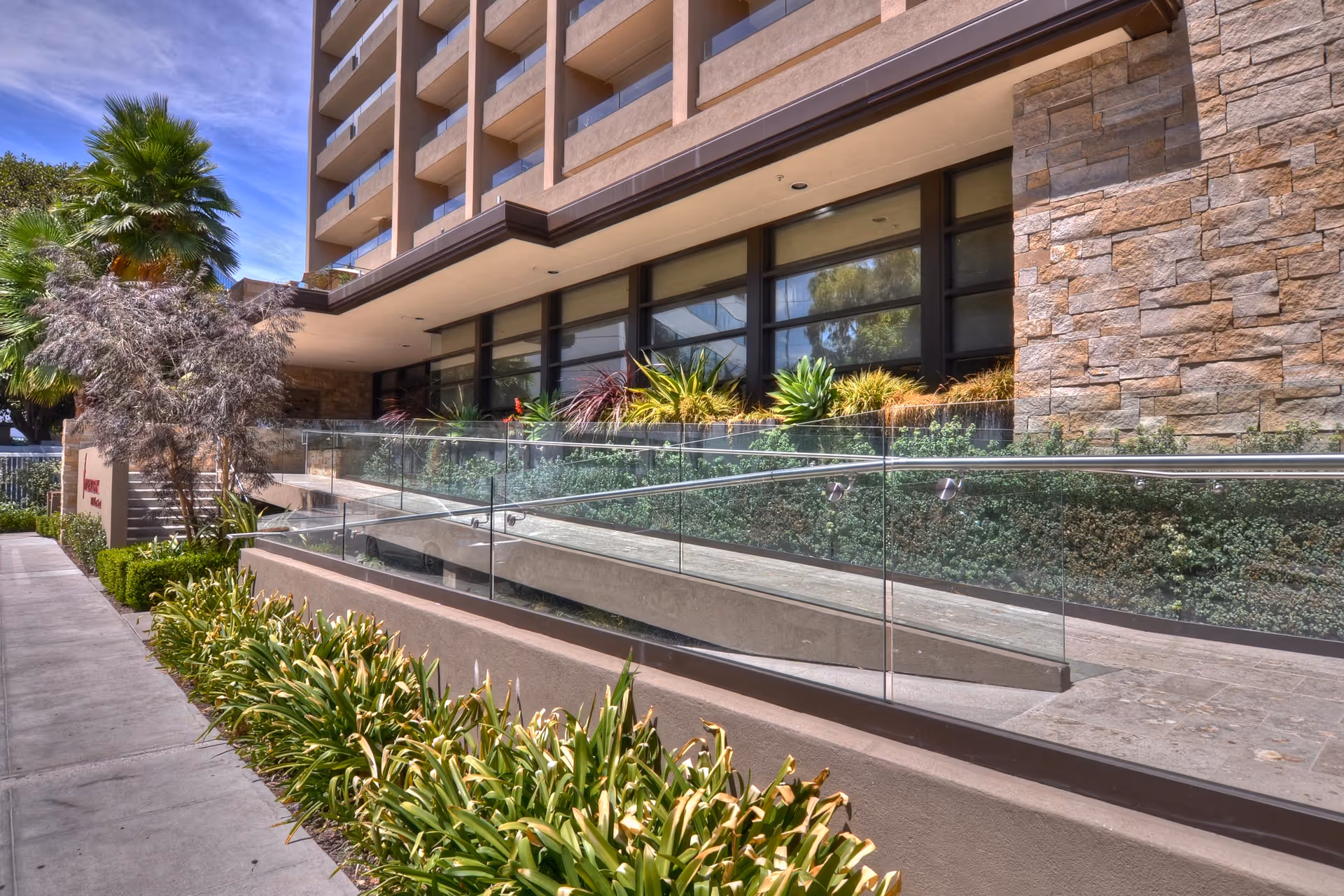 Modern senior living building exterior with glass railing, ramp, and landscaped walkway.