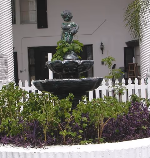 A decorative tiered fountain surrounded by white fencing and greenery in a courtyard.