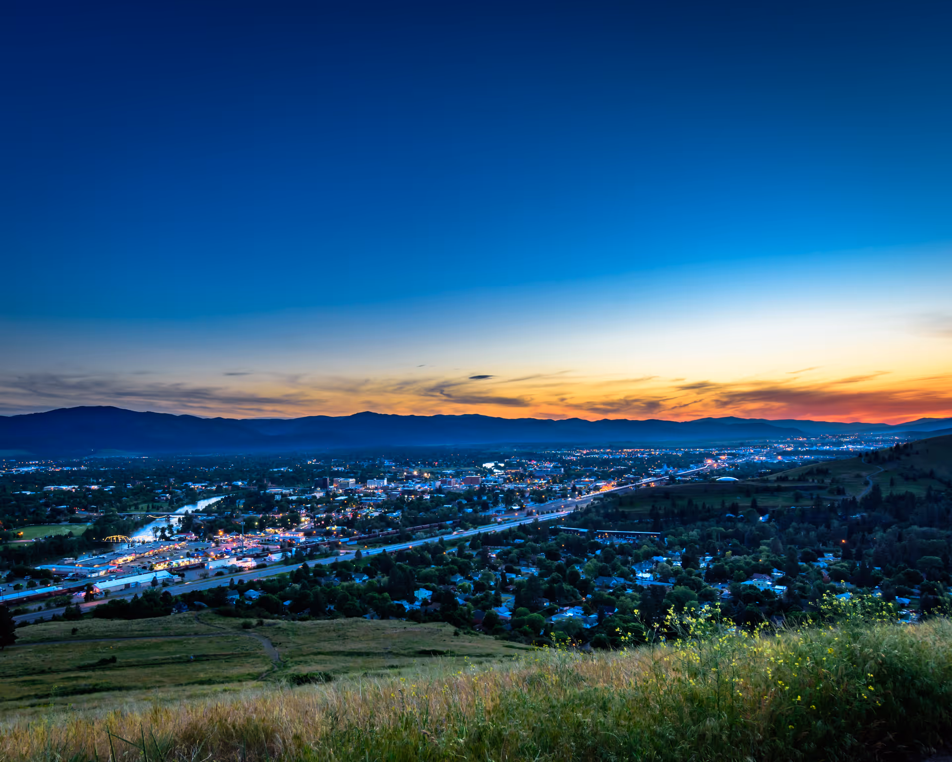 Sunset over a valley town with hills and distant mountains at dusk.