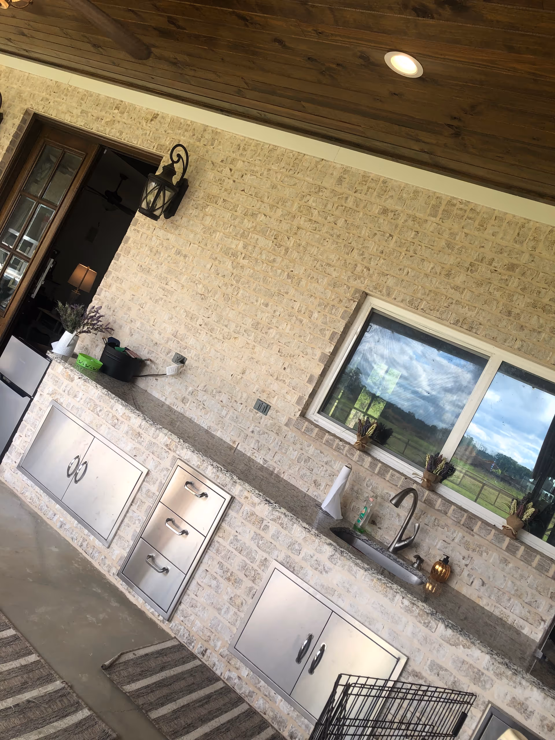 Outdoor kitchen area with brick walls, granite counter, sink, cabinets, and window