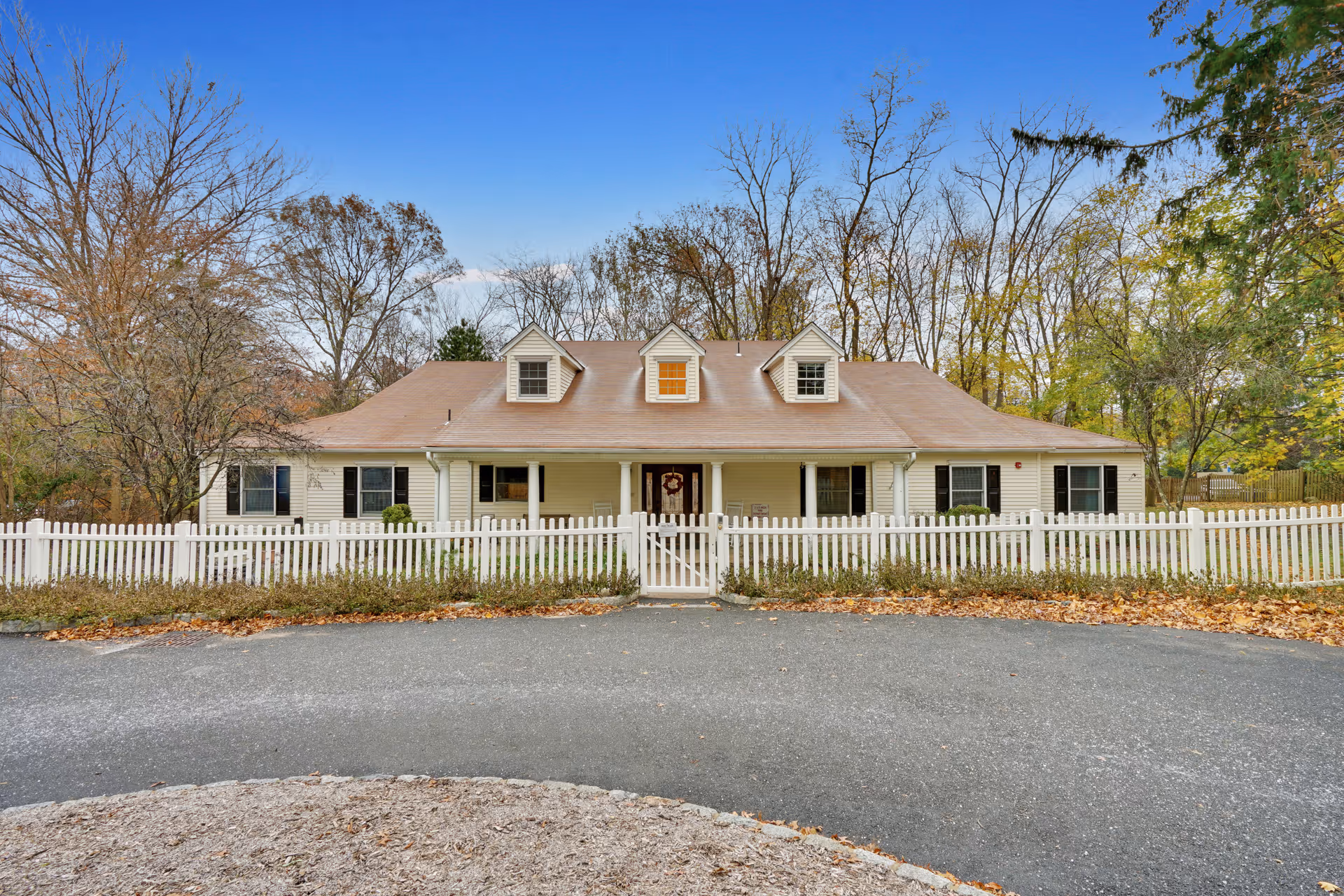Front exterior view of a single-story house with a beige exterior, three dormer windows on the roof, and a white picket fence surrounding the property. The house is set against a backdrop of leafless trees and a clear blue sky.