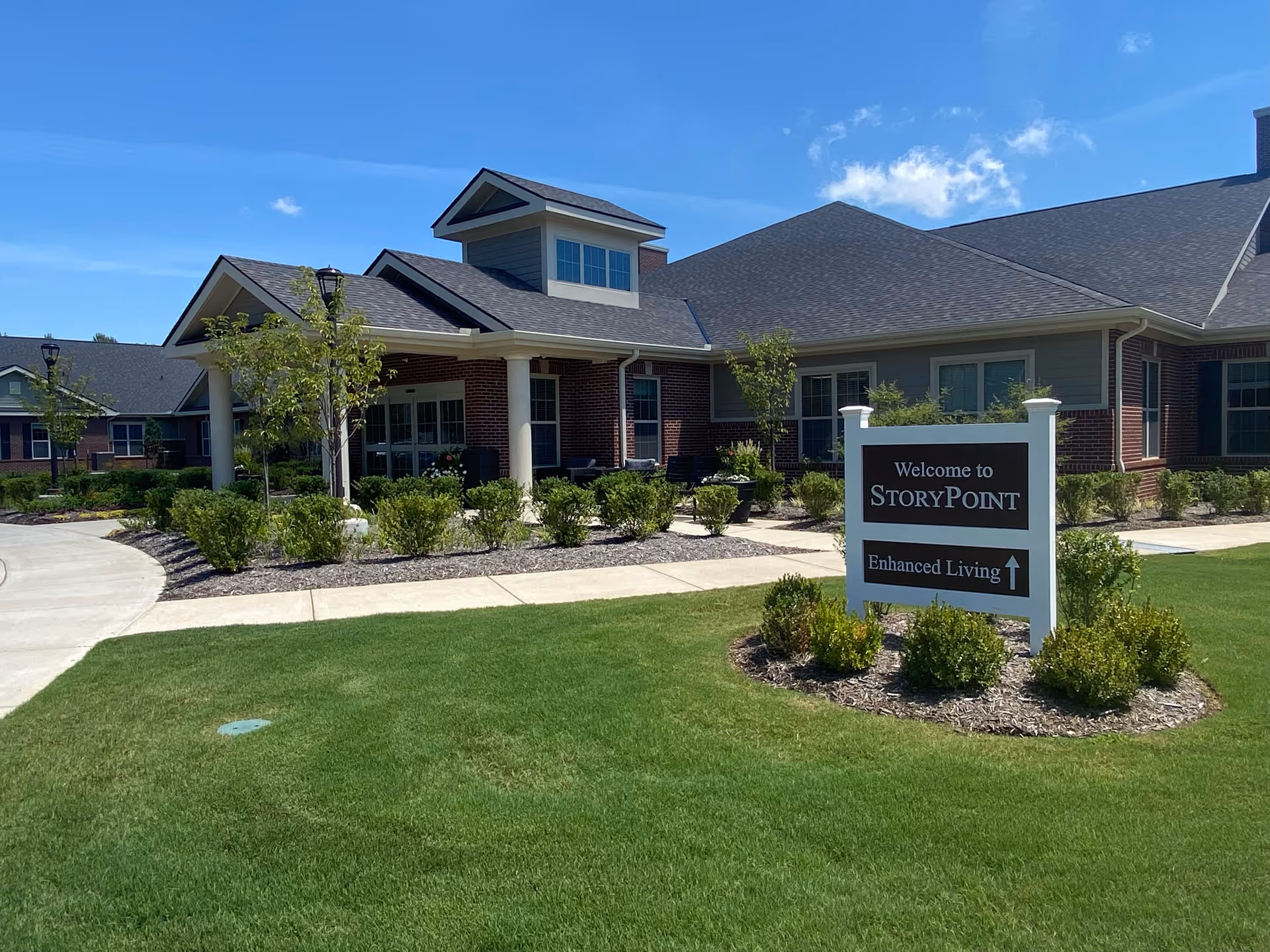 Exterior view of a single-story senior living facility building with a covered entrance supported by white columns, surrounded by neatly trimmed bushes and green lawn under a clear blue sky. A sign in front reads 'Welcome to StoryPoint Enhanced Living' with an arrow pointing forward.