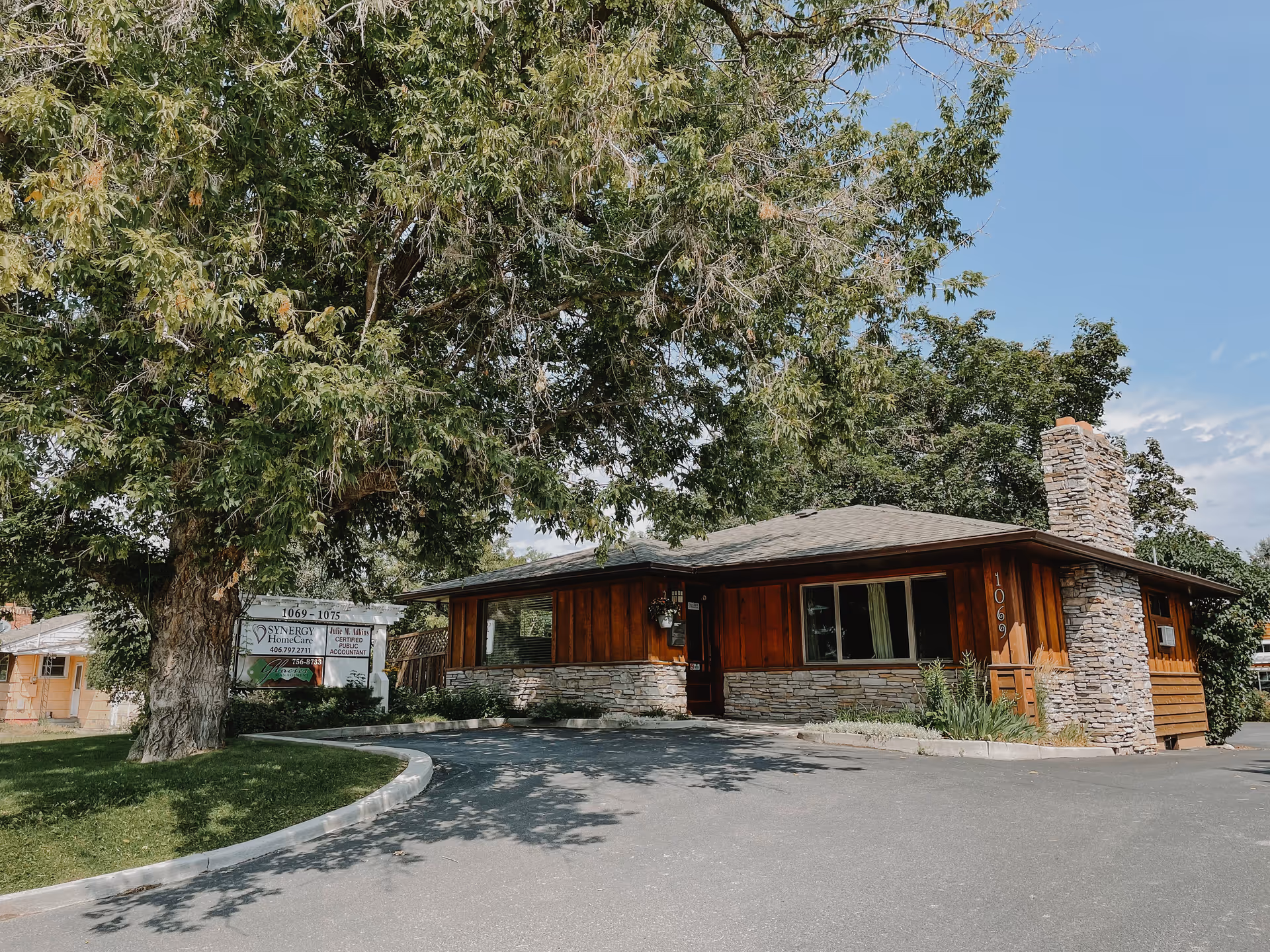 Front view of Synergy HomeCare of Flathead Valley facility, wooden exterior, stone chimney, large tree nearby