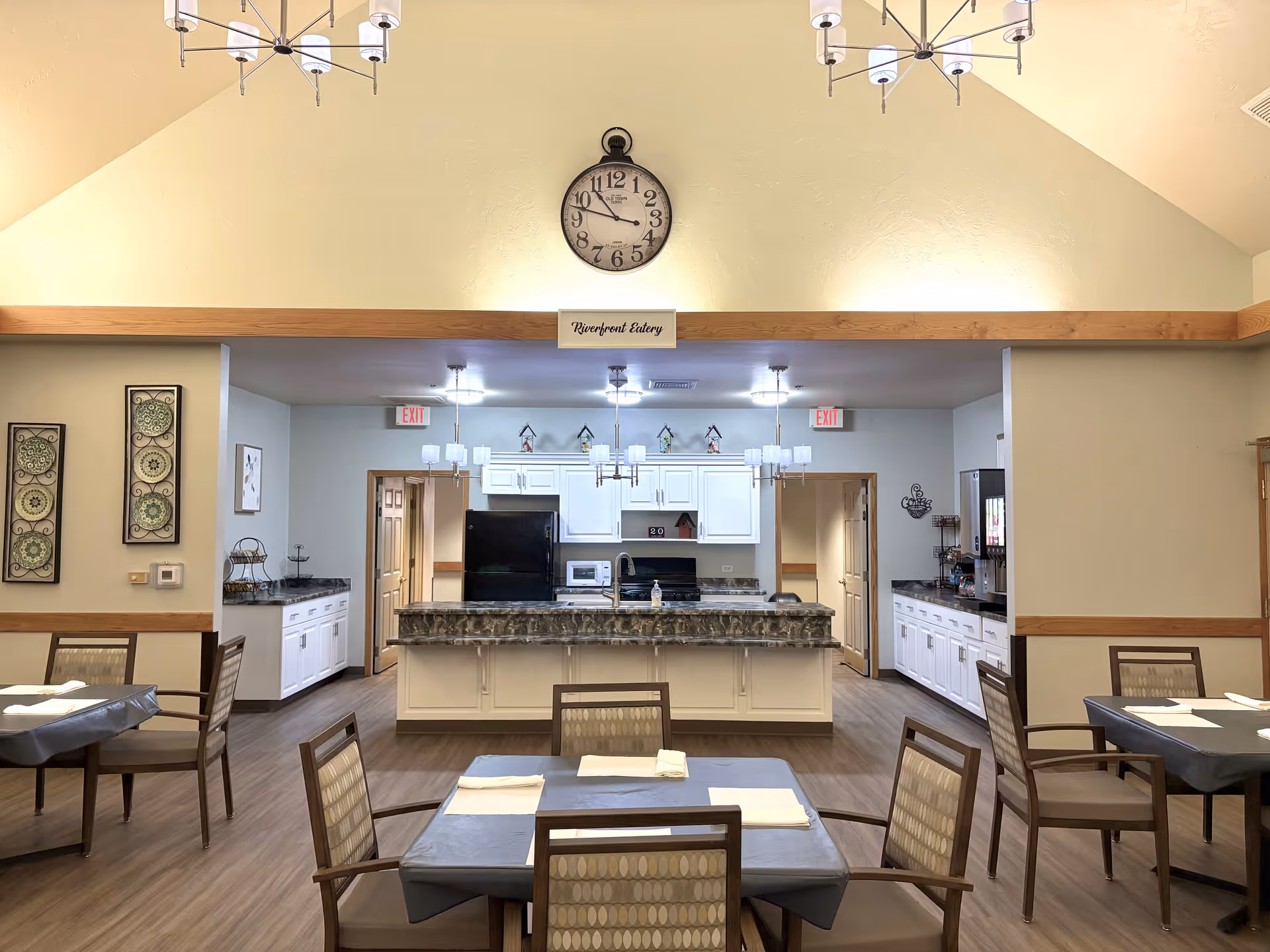 Senior living dining room with tables, chairs, and a central kitchen island beneath a clock.