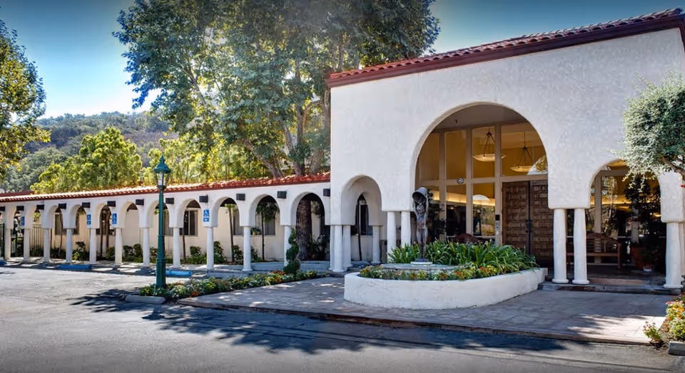 Exterior front entrance of a Mediterranean-style building with arched colonnade, columns, central planter and parking spaces.