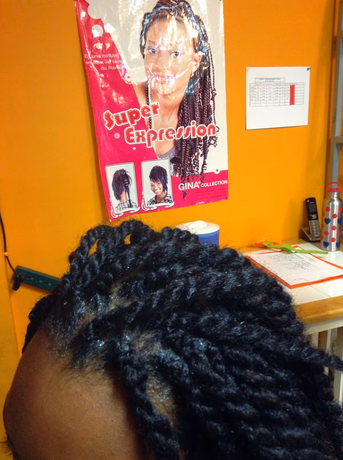Close-up of braided hair in foreground with orange wall, poster, and desk items.