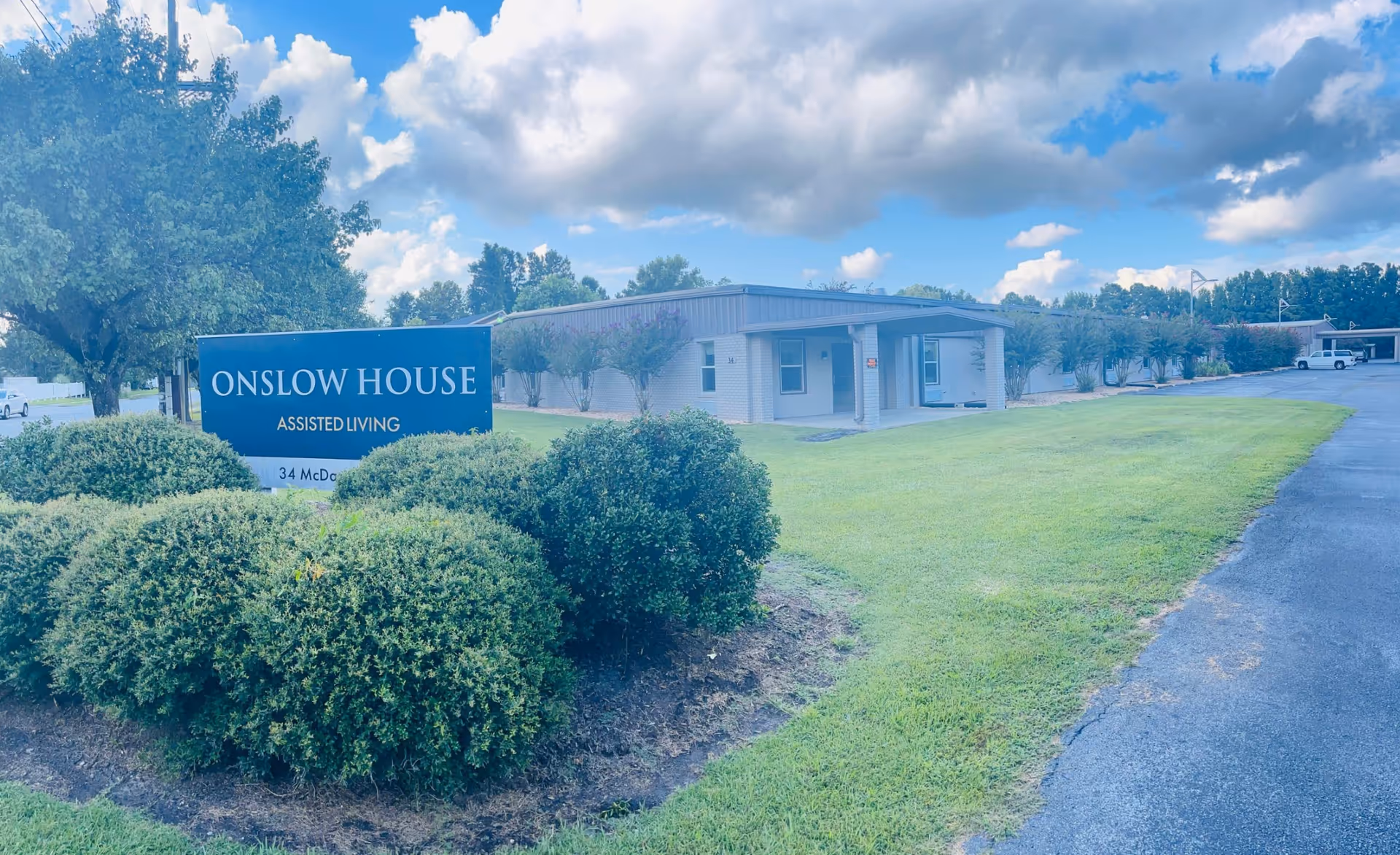 Exterior view of Onslow House Assisted Living facility with a blue sign in front surrounded by bushes and a tree. The building is a single-story structure with a covered entrance, set against a partly cloudy sky.