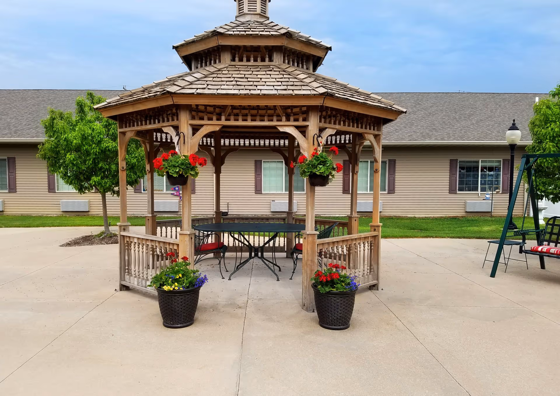A wooden gazebo with a shingled roof situated on a concrete patio. Inside the gazebo is a black metal table with matching chairs that have red cushions. Hanging flower pots with red flowers are attached to the gazebo, and two large flower pots with red and purple flowers are placed at the entrance. In the background, there is a beige building with windows and green grass, along with a swing set and a lamppost on the right side.