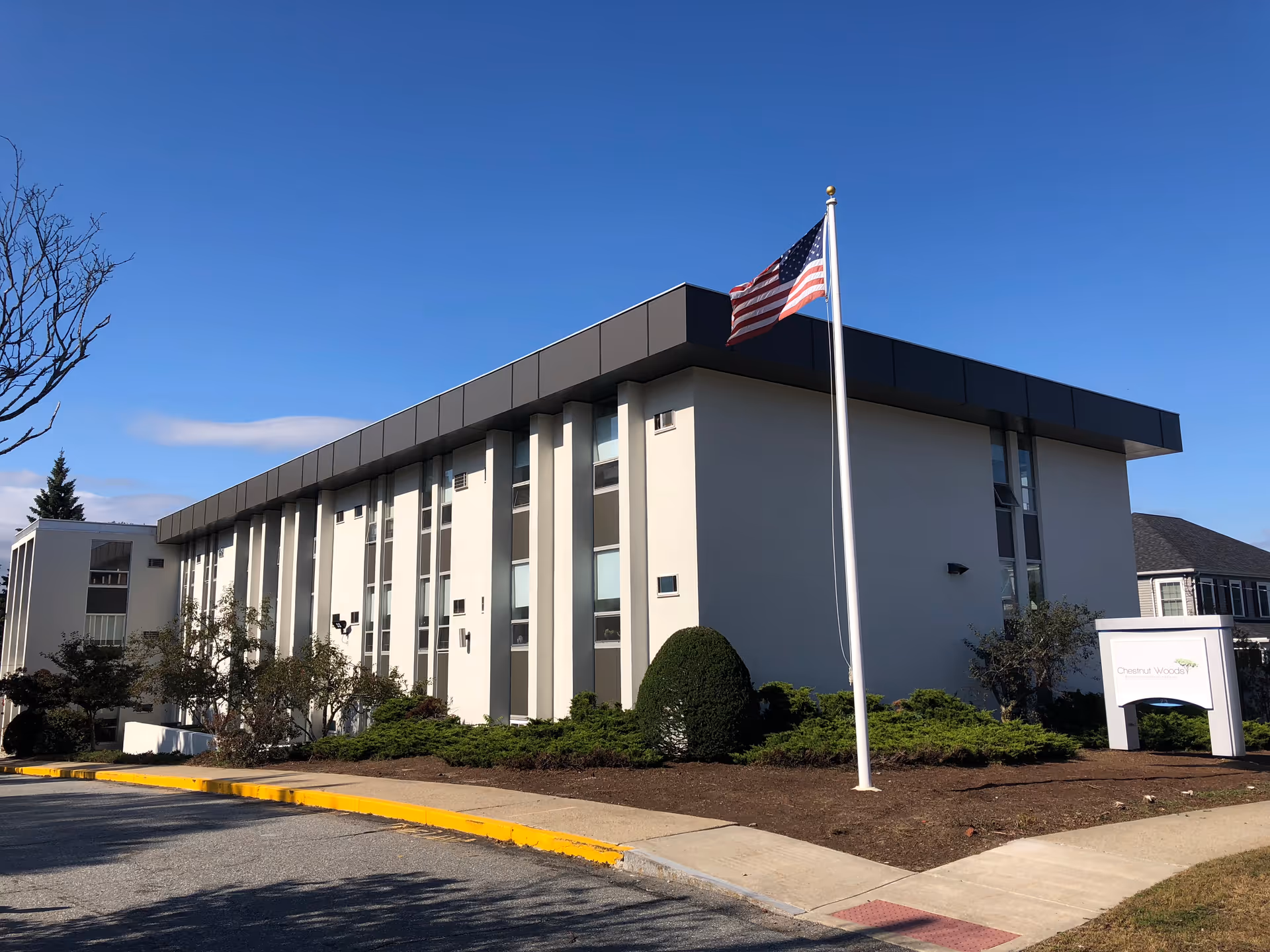 Exterior view of a multi-story building with white and gray walls, several windows, and an American flag on a flagpole in front. There are bushes and a sidewalk surrounding the building, and a sign near the entrance reads 'Chestnut Woods Rehabilitation & Healthcare Center.' The sky is clear and blue.