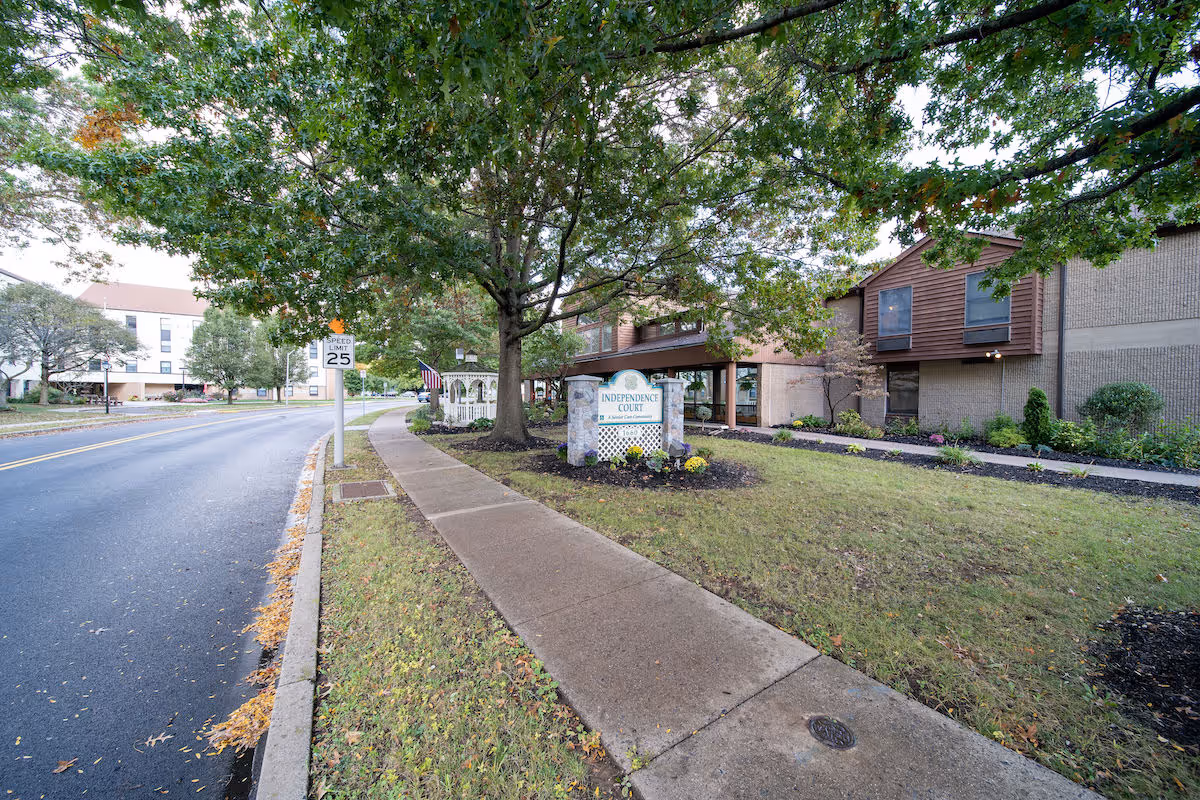 Sidewalk leading to the entrance of Independence Court of Quakertown, a senior living facility. The building is surrounded by grass, trees, and landscaped flower beds. A speed limit sign of 25 mph is visible along the adjacent road.