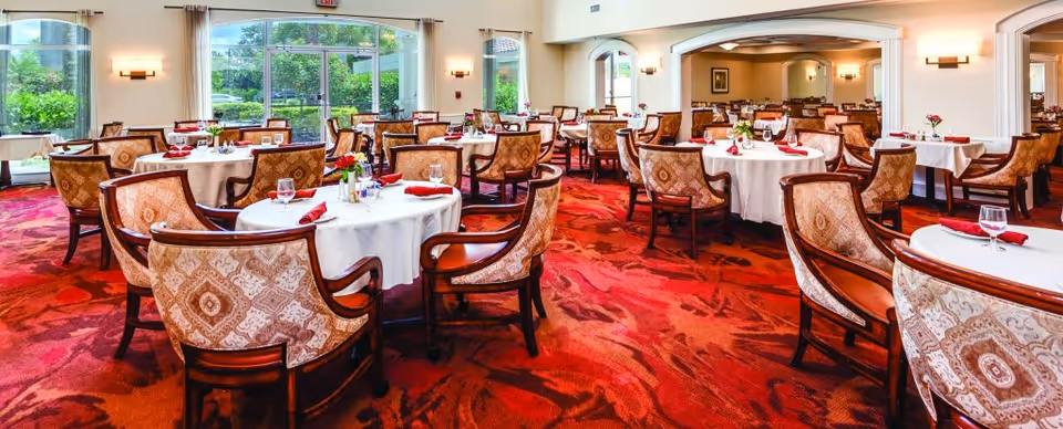 Spacious dining room with round tables set with white tablecloths and patterned chairs on a red carpet.