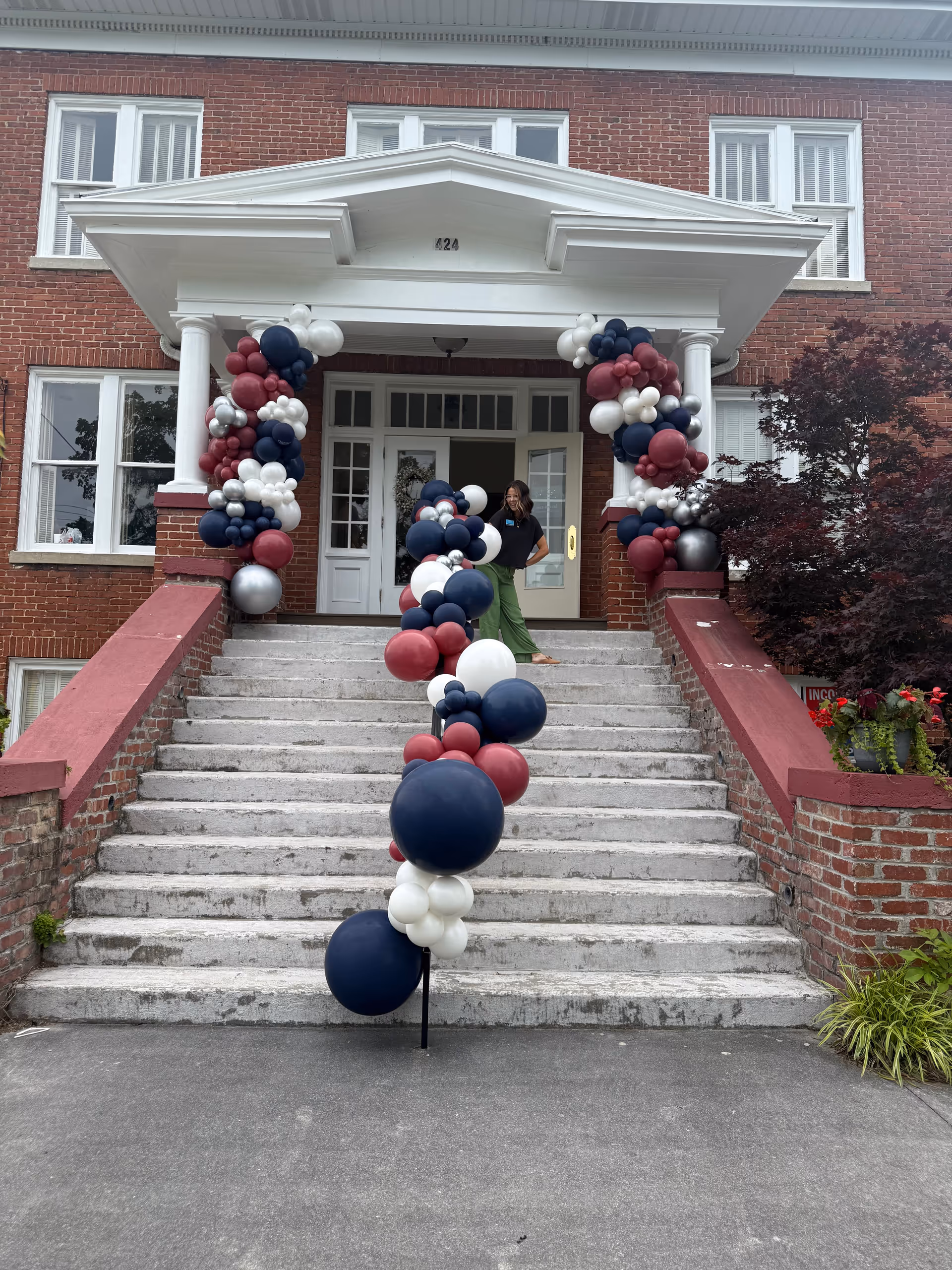 Brick building entrance of SYNERGY HomeCare decorated with red, white, and navy balloons along stairs.