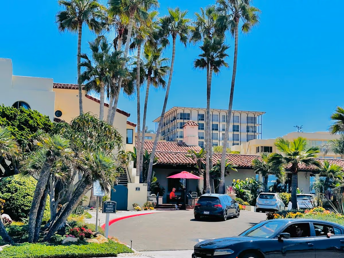 Entrance and front facade of a Mediterranean-style retirement community with palm trees, a driveway with parked cars, and a red curb under a clear blue sky.