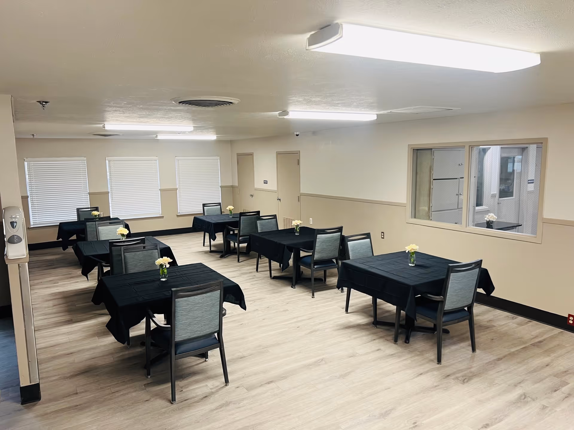 A dining room with six tables covered in black tablecloths, each table has two chairs and a small vase with white flowers. The room has light wood flooring, beige walls with a chair rail, three windows with closed blinds on one side, and a window looking into another room on the right wall. The ceiling has fluorescent lights.