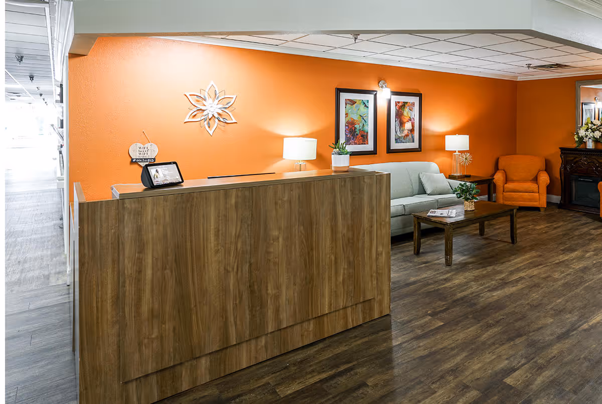 Reception area with a wooden front desk, an orange accent wall behind it, two framed colorful artworks, a gray sofa, an orange armchair, a wooden coffee table with a plant and magazines, two table lamps, and a decorative fireplace with flowers on the right side.