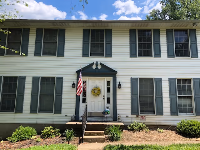 Two-story white building with dark shutters, front steps, flag, wreath, and landscaping.