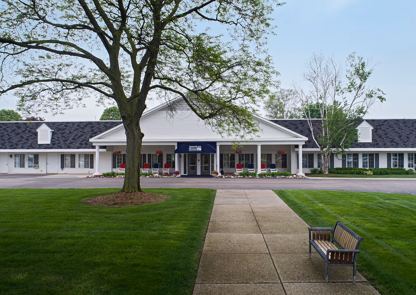 Front exterior of American House Dearborn Heights senior living facility, featuring entrance, greenery, and a bench.