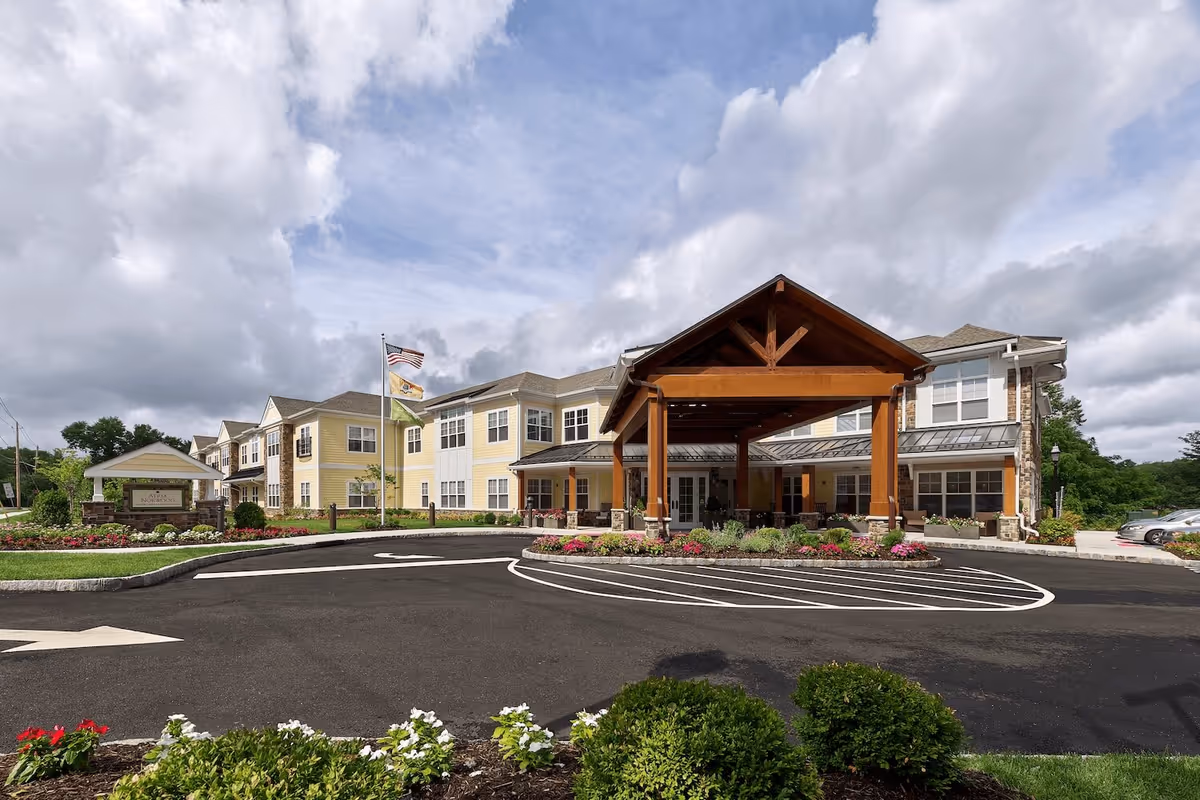 Exterior view of a two-story assisted living facility with a large covered entrance supported by wooden beams. The building is painted yellow with white trim and has multiple windows. There are landscaped flower beds and bushes in front, a paved driveway with directional arrows, and three flags flying on flagpoles near the entrance. The sky is partly cloudy.