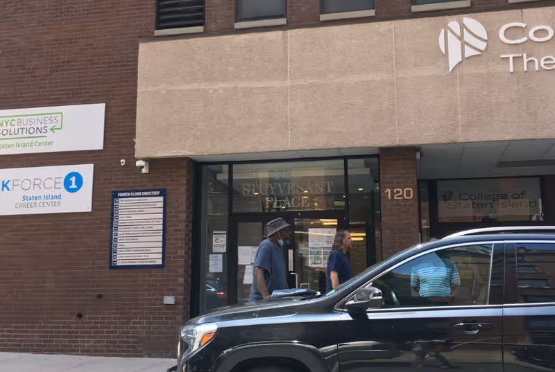 Exterior of a brick building with glass doors and people walking beside a parked SUV.