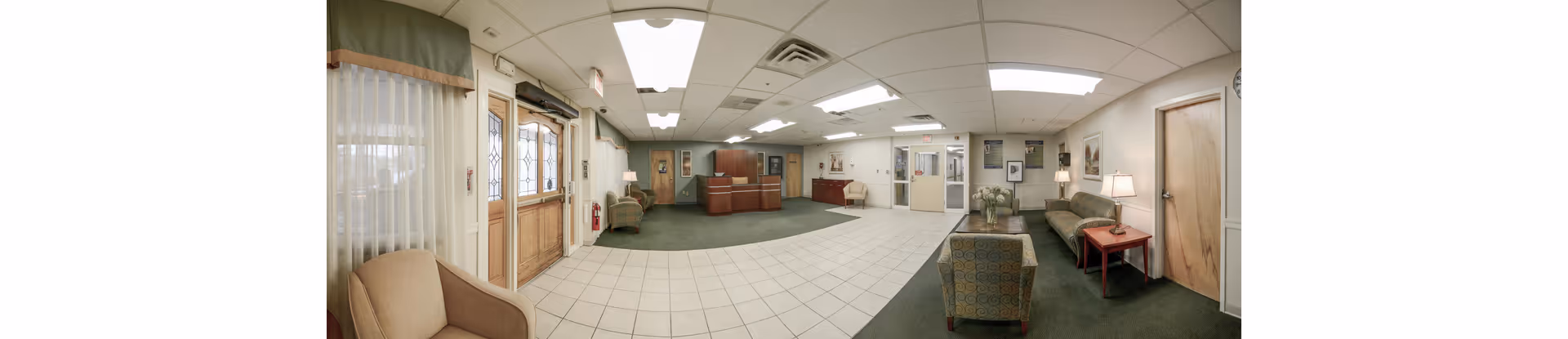 Wide panoramic view of a senior living facility's lounge with couches, chairs, and a reception area.