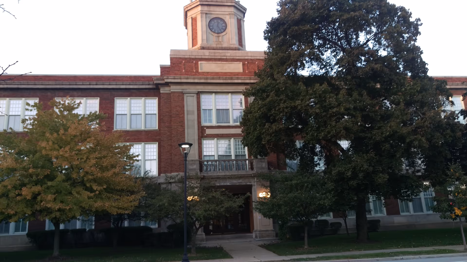 Brick building facade with clock tower overlooks trees and a walkway leading to the entrance.