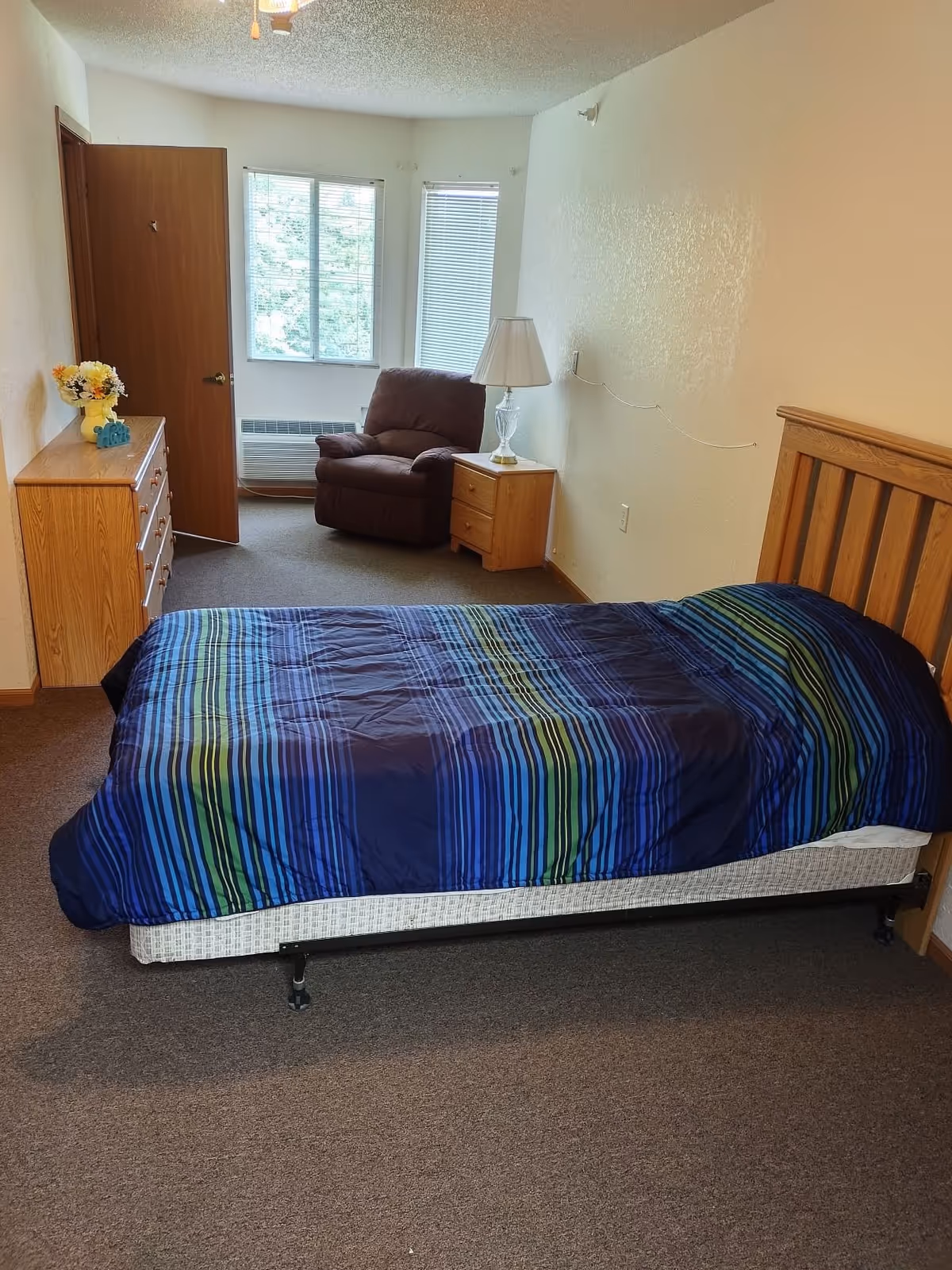 A bedroom with a single bed covered in a blue and green striped comforter. There is a wooden headboard, a wooden dresser with a vase of flowers on top, a brown armchair near two windows, and a wooden nightstand with a lamp. The walls are light-colored and the floor is carpeted.