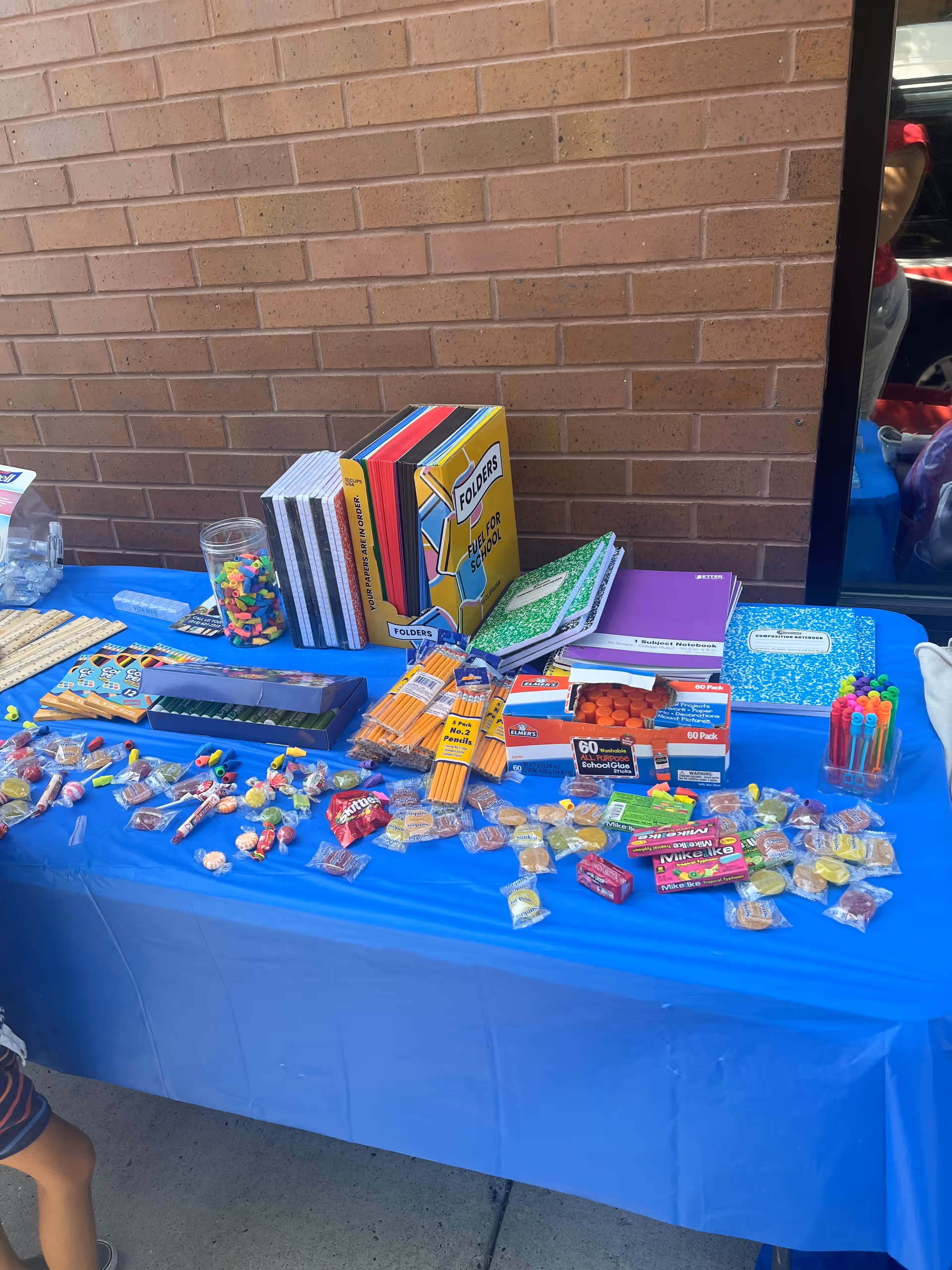 A blue tablecloth covers a table outside, displaying assorted school supplies and candy.