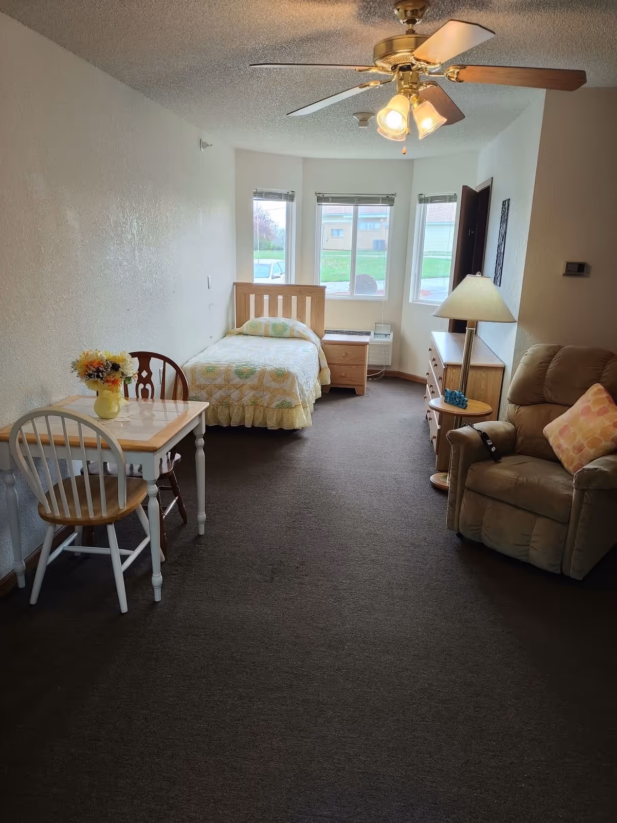 Studio-style senior living room with a single bed, recliner, small dining table and dresser beneath bay windows.