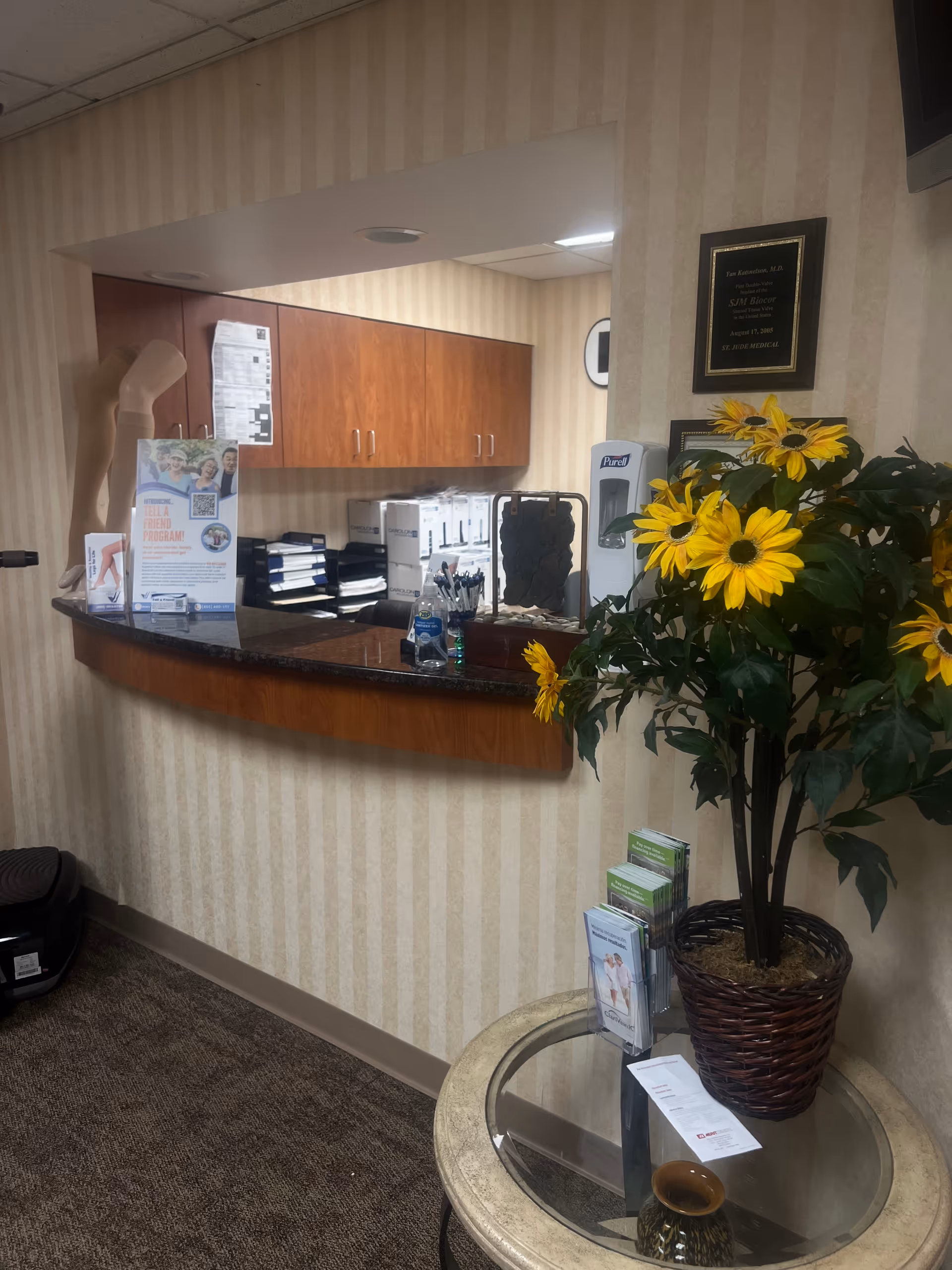 Reception desk with wood cabinets, a sunflower plant, brochures, and a round glass table.