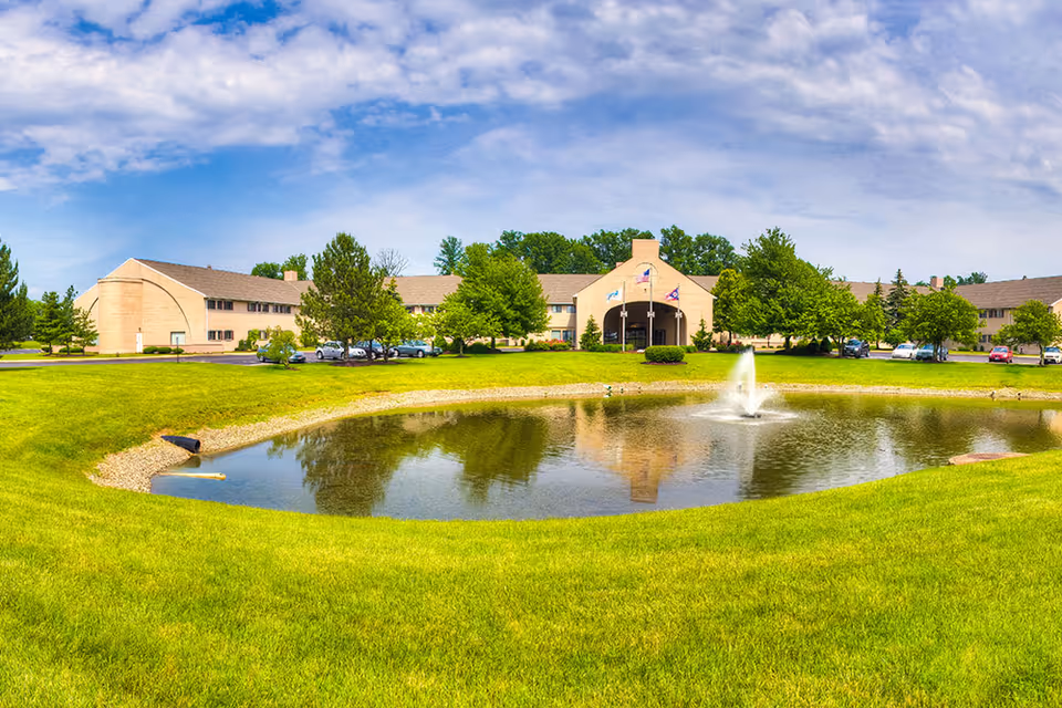 Wide exterior view of The Abbewood senior living facility showing a large building with multiple windows, surrounded by green trees and a well-maintained lawn. In the foreground, there is a pond with a water fountain in the center, reflecting the building and sky above. Several cars are parked near the building under a partly cloudy sky.