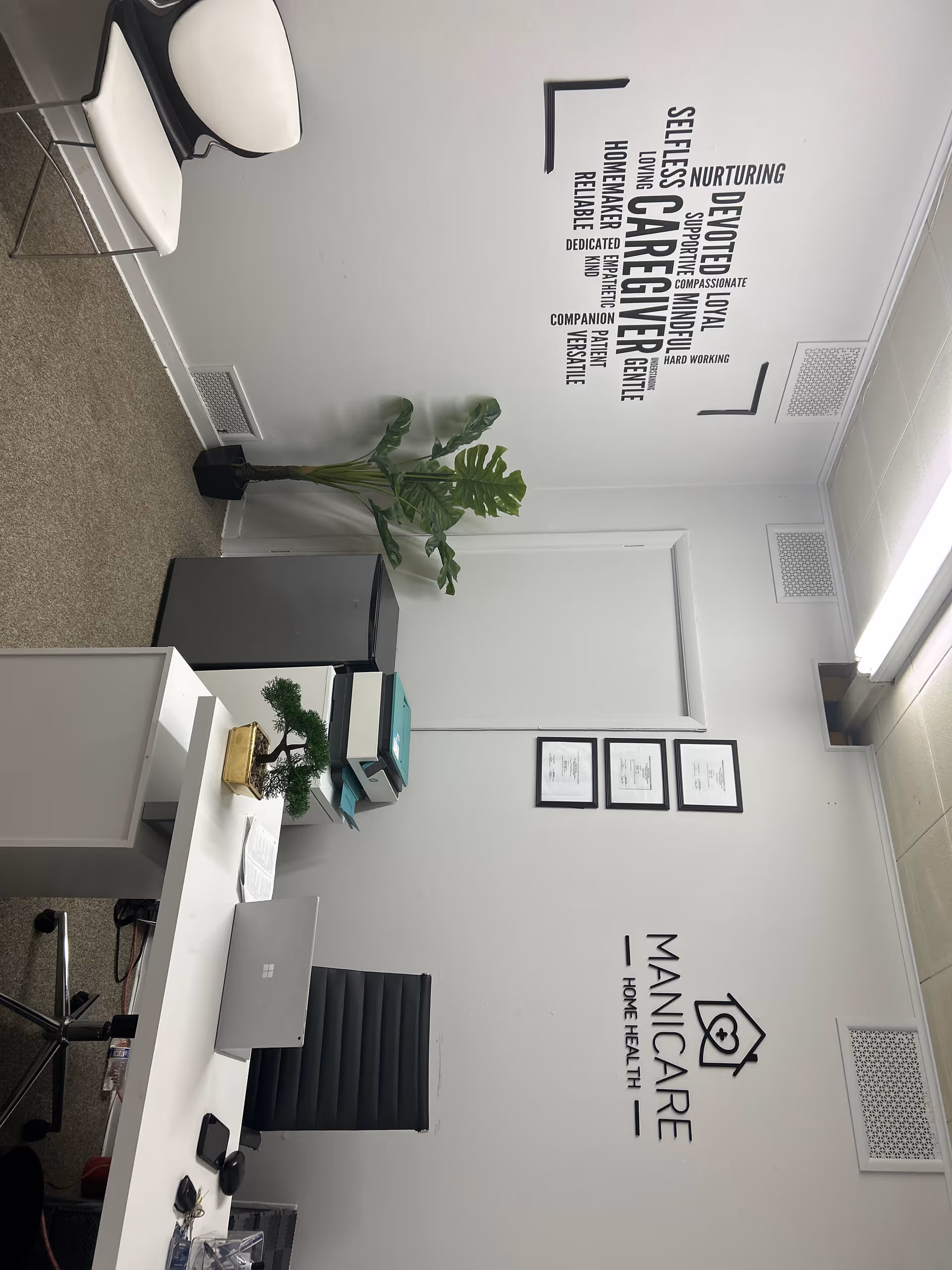 Office reception area of Manicare Home Health with desk, chair, plant, and certificates.