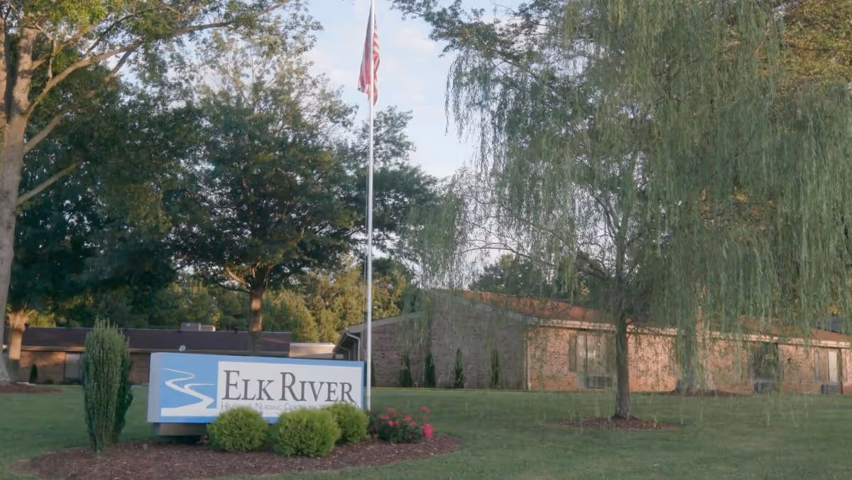 Exterior view of Elk River Health and Nursing Center of Winchester showing a brick building surrounded by trees and greenery, with a sign in the foreground displaying the facility's name and an American flag on a flagpole behind the sign.