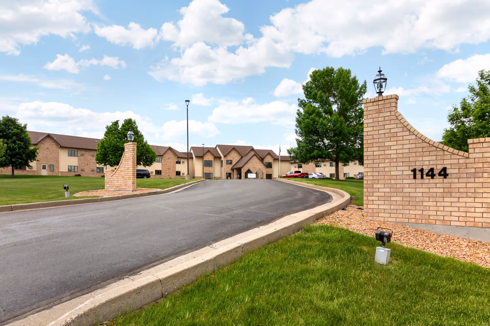 View of the front exterior of Hay Creek Lodge Senior Living facility with a paved driveway leading to the entrance. The building is two stories with a combination of brick and beige siding. There are green lawns, trees, and a brick sign with the number 1144 near the driveway entrance under a partly cloudy sky.