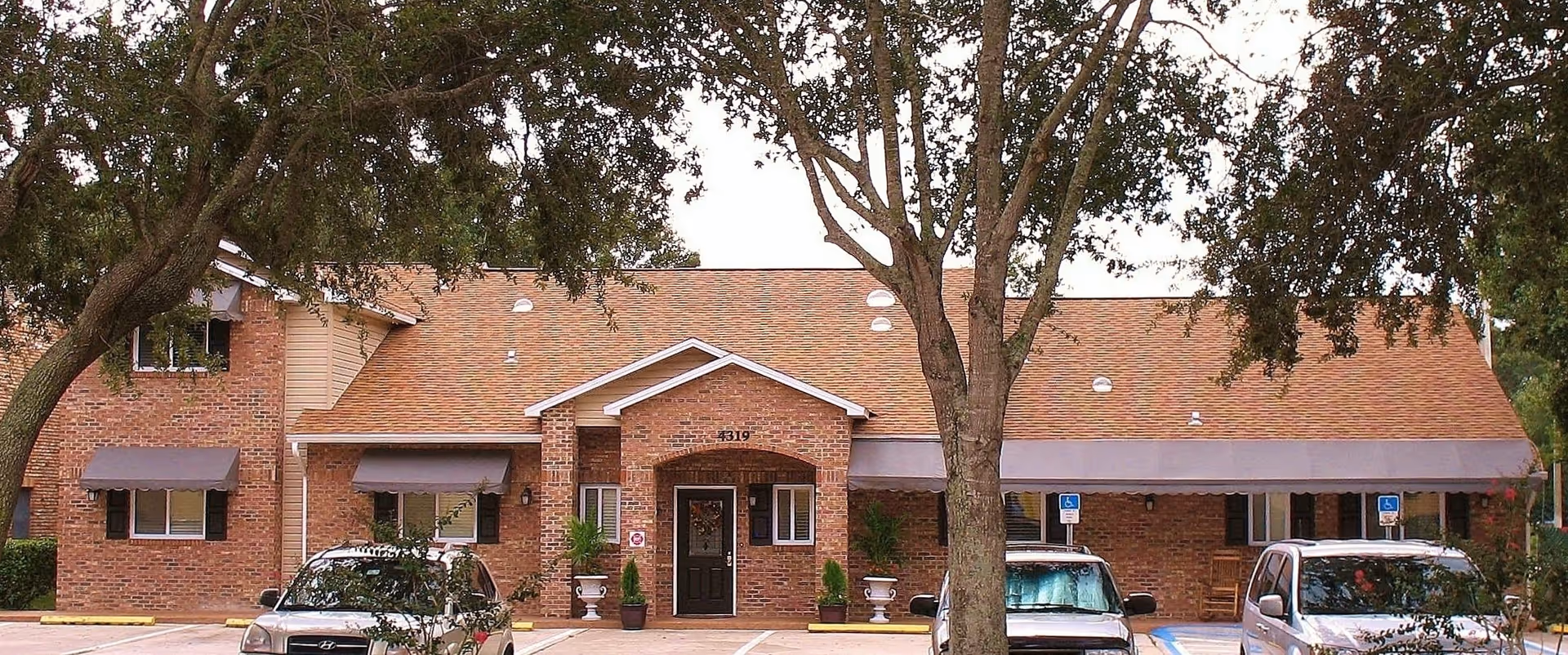 Front exterior of a brick residential-style building with a central entrance, awnings, trees, and parked cars.