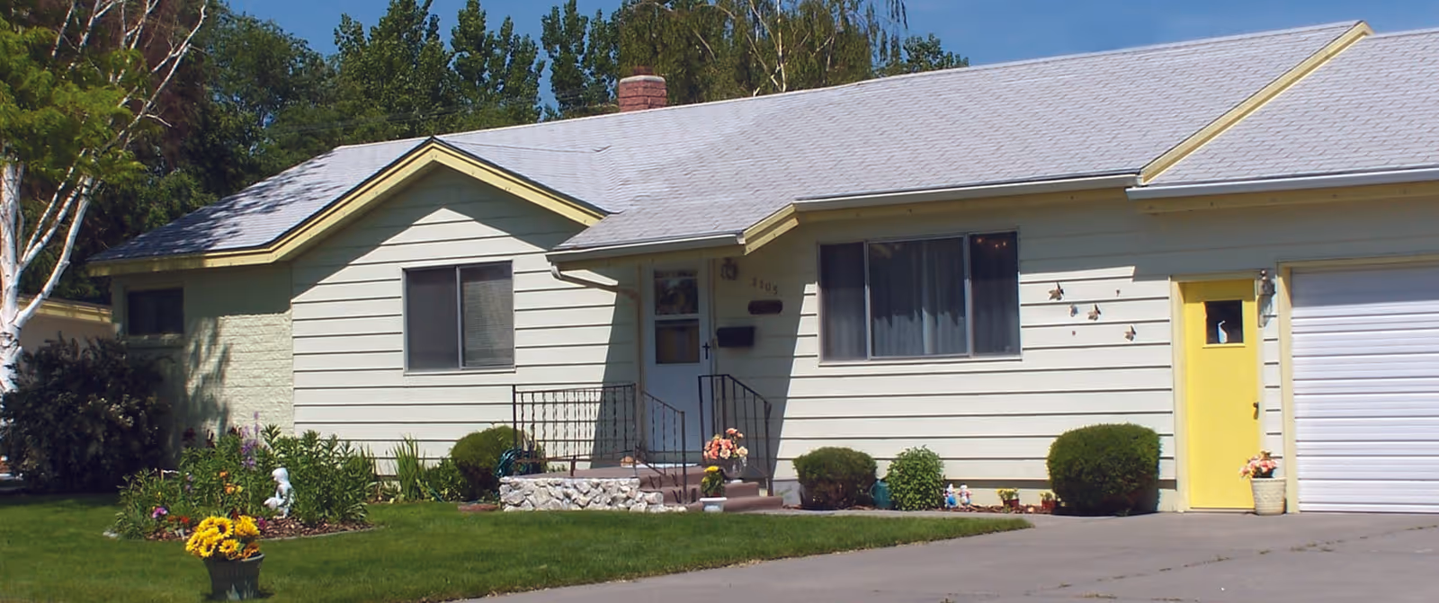 Single-story pale yellow house with a front lawn, flowerbeds, a yellow entry door, and an attached garage.