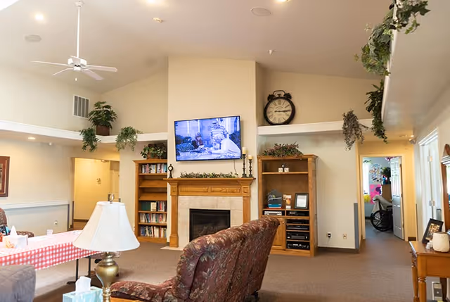 A cozy living room area in Autumn Haven Assisted Living featuring a fireplace with a mounted TV above it, flanked by wooden bookshelves filled with books and decorative items. There is a patterned armchair in the foreground, a table with a red and white checkered tablecloth to the left, and ceiling fans and hanging plants adding to the homey atmosphere.