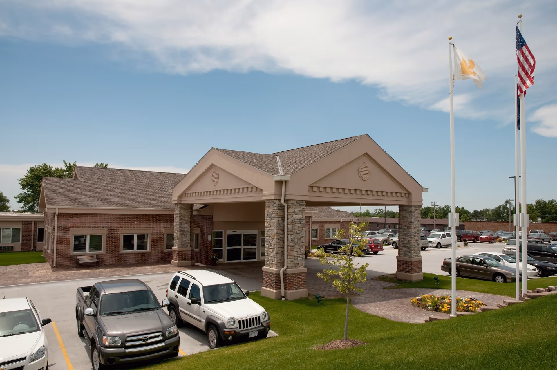 Exterior view of Life Care Center of Elkhorn showing the main entrance with a covered drop-off area supported by stone columns. Several cars are parked in the parking lot, and two flagpoles with flags are visible on the right side. The sky is partly cloudy.