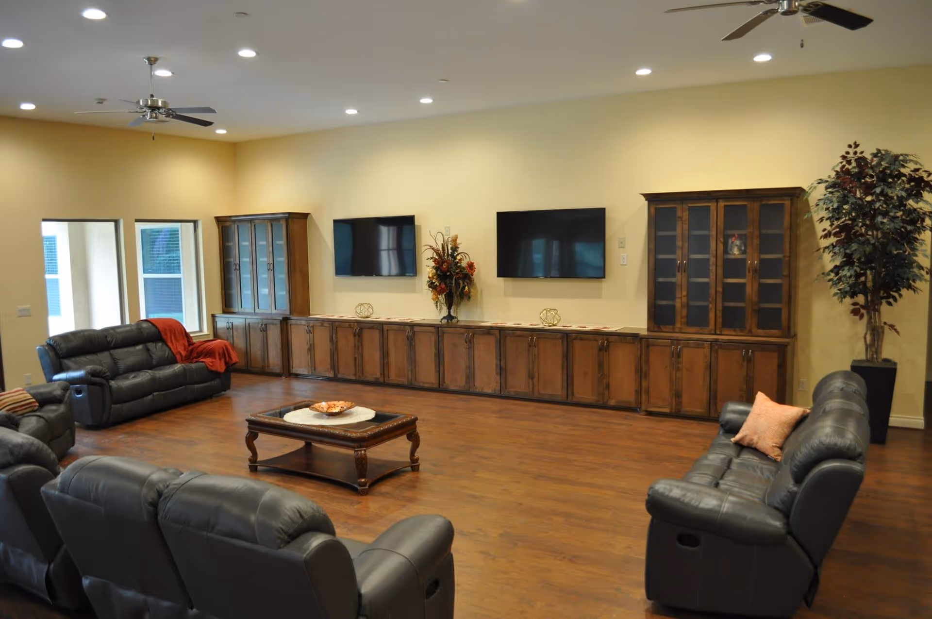 A spacious living room with multiple black leather recliner sofas arranged around a wooden coffee table. The room features wooden flooring, two flat-screen TVs mounted on a beige wall, and long wooden cabinets beneath the TVs. There are two tall wooden display cabinets on either side of the TVs and a large potted plant in the corner. Ceiling fans with lights are mounted on the ceiling.