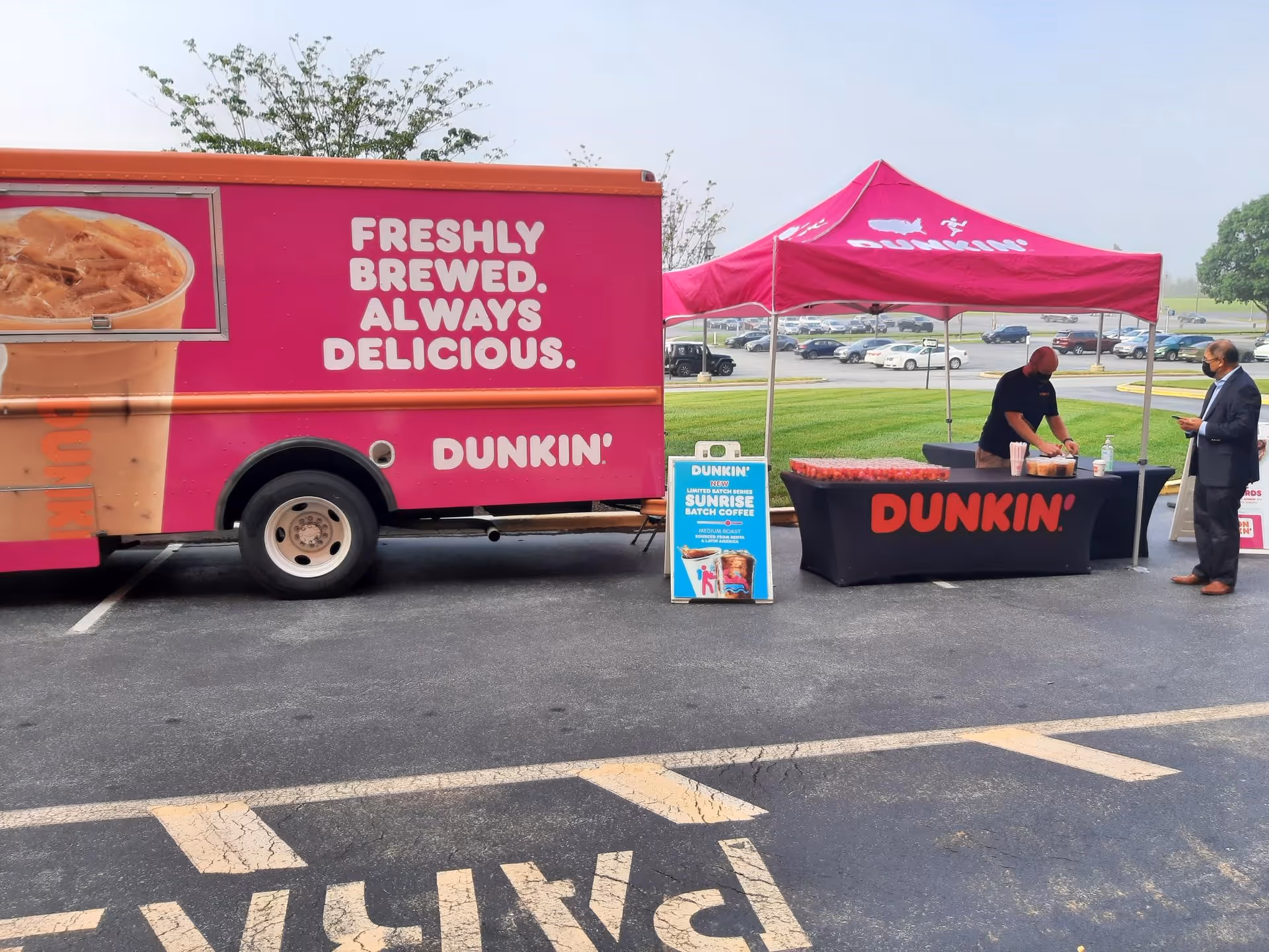 A pink Dunkin' truck and matching tent set up in a parking lot promoting coffee.