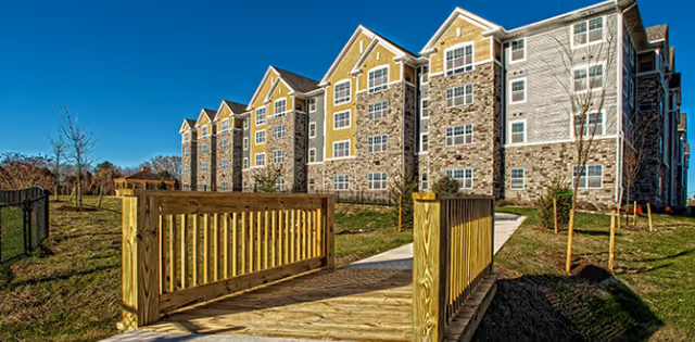 Exterior view of a multi-story senior living residence with stone facade and wooden bridge
