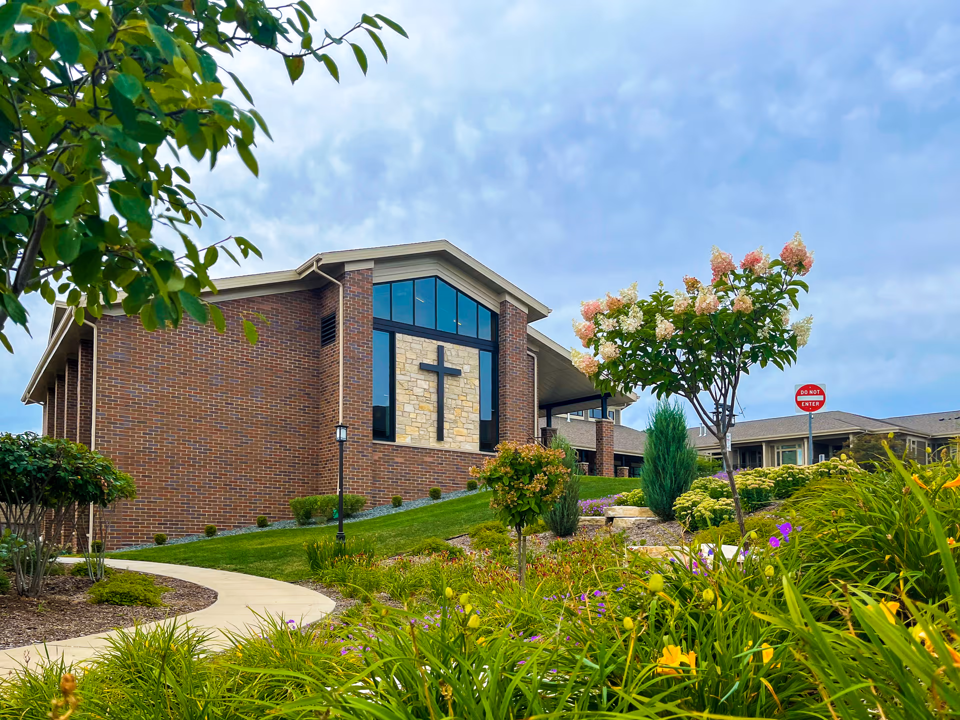 Exterior view of a brick building with large windows and a cross mounted on a stone section. The building is surrounded by a well-maintained garden with various plants, flowers, and a curved sidewalk. A 'Do Not Enter' sign is visible in the background under a partly cloudy sky.