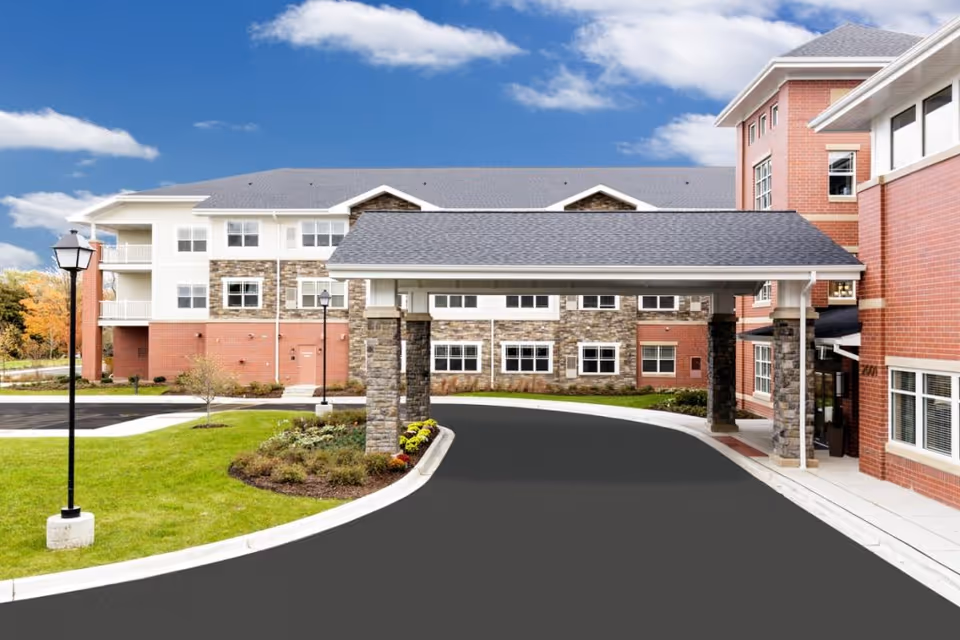 Exterior view of Clarendale of Algonquin senior living facility showing a covered driveway entrance with stone pillars, a multi-story building with brick and stone facade, green lawn, and a clear blue sky with some clouds.