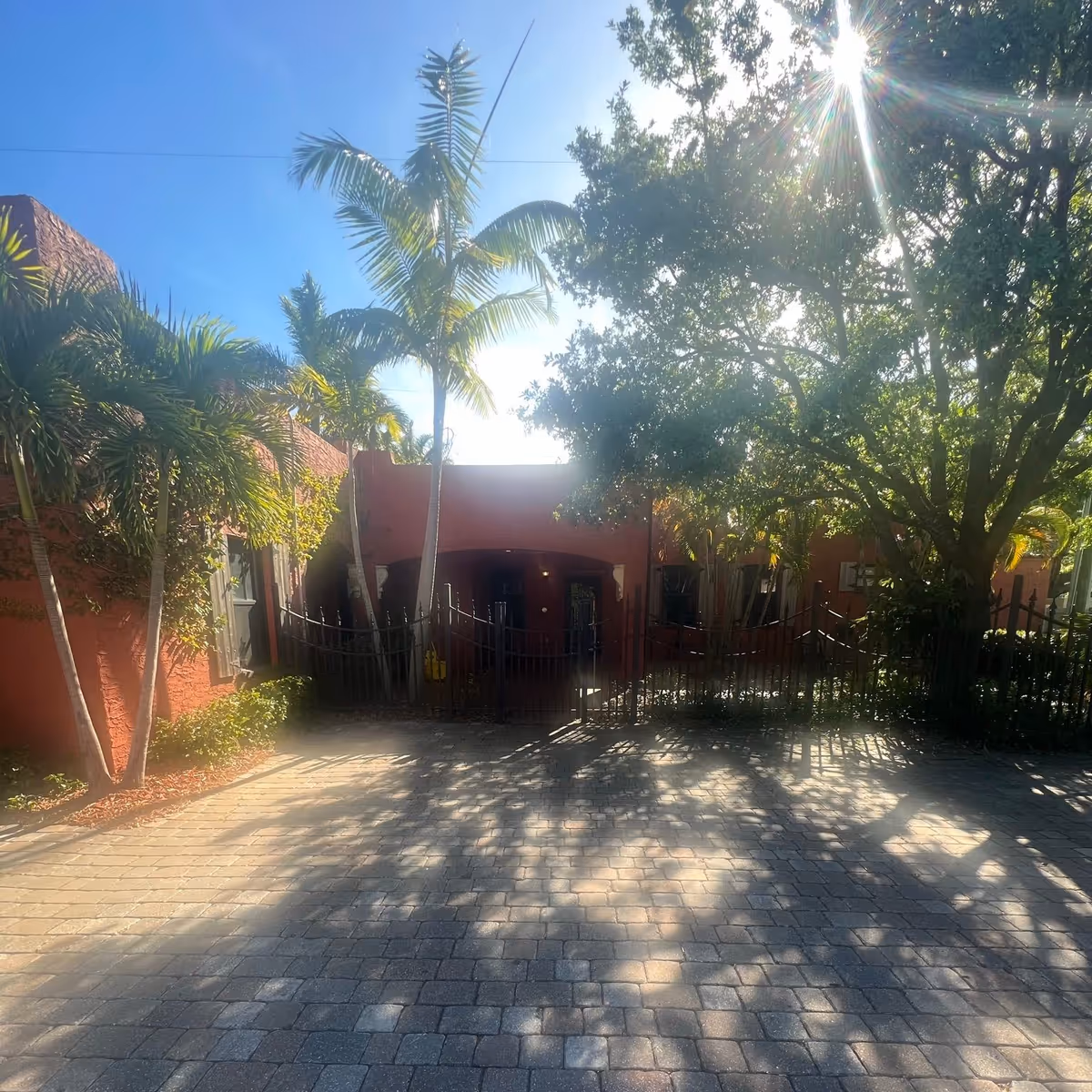 Sunlit entrance to a red brick senior living facility, with palm trees and a gated walkway.