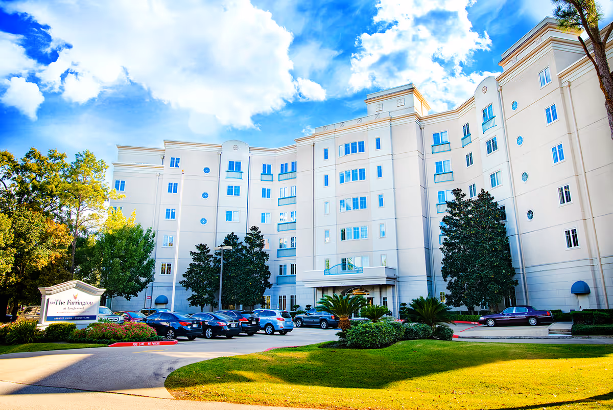 Front view of a large multi-story cream-colored senior living building with parked cars and landscaped grounds under a bright blue sky.